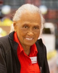 An elderly woman with gray hair pulled back smiles warmly, wearing a red collared shirt, black jacket, and name tag in a market.
