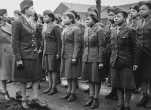 Women in WWII military uniforms stand in formation outdoors, listening as a uniformed woman inspects them.