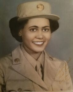 A young woman in a khaki military uniform and cap smiles at the camera; sepia photo with a handwritten note in the corner.