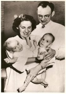 A smiling woman cradles two conjoined infants, while a man supports one baby's back. All four wear light-colored clothes.