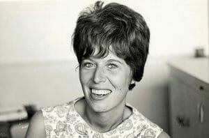 Black and white photo of a young woman with short dark hair, smiling, in a sleeveless top. Plain wall and cabinet in background. It appears to be a photo from the mid-20th century.