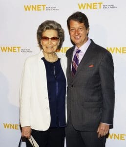 An older woman in sunglasses and a white jacket stands with a suited man, both smiling before a WNET New York Public Media banner.