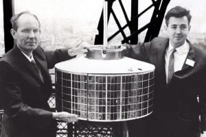 Two men in suits stand on either side of a cylindrical satellite with solar panels, holding it for display. Metal structure and cityscape are visible in the background. The image is in black and white.