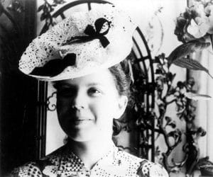 A young woman in a patterned dress smiles softly, wearing a large, decorative hat with lace and a ribbon. She stands indoors near an ornate metal gate and floral decorations. The photo is in black and white.