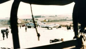 Military airbase with people by two aircraft on tarmac; photo taken from behind a dark object, buildings and hills in background.