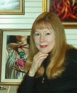 A woman with long, straight light brown hair and bangs poses indoors, her chin resting on her hand. Framed art hangs behind her.