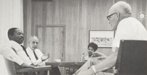 Four people in an office having a serious discussion; one man gestures, two men listen, and a woman sits at the table. Wood-paneled walls.