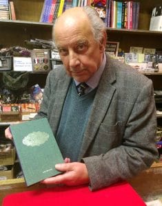 An older man in a gray suit and blue sweater stands in a bookstore, holding a green hardcover book and looking at the camera. Shelves of books and various items are visible in the background.