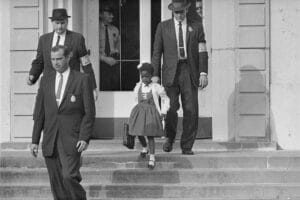 A young Black girl in a dress with a school bag is escorted by four suited men as she walks down the steps of a building.
