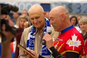 Two bald men stand together; one reads from a plaque into a mic, the other smiles. A crowd surrounds them; left man wears blue-white scarf.