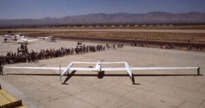 A large white experimental aircraft with a long wingspan sits on a runway apron, crowds nearby, and mountains in the background.