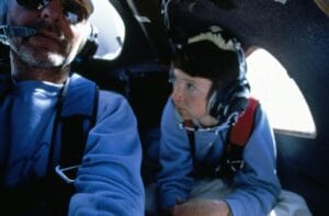 A man and woman in headsets and blue clothes sit in a cockpit; the woman looks at the man as sunlight streams through a window.