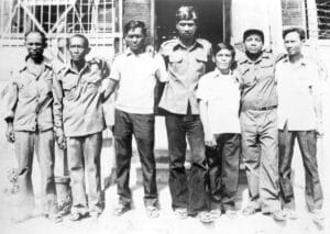 Seven men stand side by side outdoors, posing for a group photo in front of a building with barred windows and a staircase, all dressed in shirts and pants. The image is in black and white.