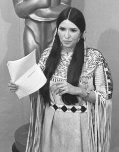 A woman in long braids and Native American attire with fringe and beads holds a paper, standing before a large Oscar statue.