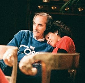 A man in a blue Cosby Show sweatshirt sits with a woman in red resting her head on his shoulder; both look relaxed, softly lit.