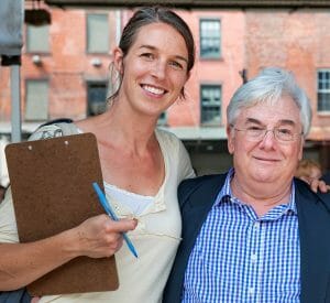 A smiling woman with a clipboard and pen stands by an older man in glasses and checkered shirt, outdoors near brick buildings.