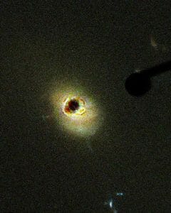 A dark circle encircled by a colorful glowing halo on black, with a rod shape extending from the upper right toward the center.