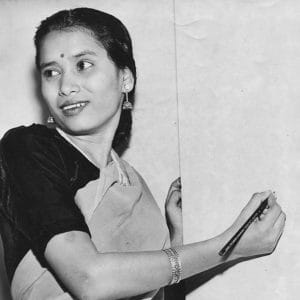 A woman in a saree with braided hair looks to the side while holding a pencil and standing in front of a blank surface, possibly a board or paper. She wears earrings, a bindi, and a bracelet.