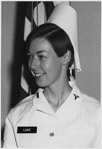 A smiling woman in a nurse’s uniform and cap stands in front of an American flag. Her name tag reads “Lane.” She has short hair and is facing slightly to the side.