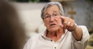 An older man with gray hair and glasses, in a light shirt, points forward while speaking. The background is softly blurred.