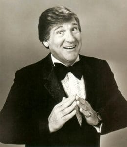 A man in a tuxedo and bow tie smiles broadly, hands clasped, posing against a plain backdrop in a black and white photo.