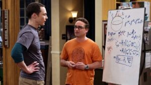 Two men chat in an apartment near a whiteboard covered with math equations and a sphere drawing. Shelves and a lamp are behind them.
