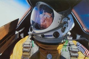 An astronaut or pilot in a yellow pressure suit and helmet sits in an aircraft cockpit, visor reflecting sky, flag patch showing.