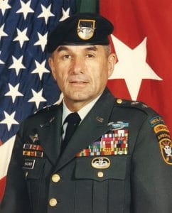A decorated military officer in dress uniform and beret stands in front of the American flag and another flag, red with a large white star. He has several badges, ribbons, and patches on his uniform.