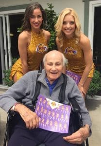 An elderly man in a wheelchair, smiling and holding a Lakers cheerleader poster, is joined by two smiling Lakers cheerleaders in gold outfits, posing outdoors in front of a building.