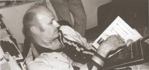 Black-and-white photo of a man sitting and looking at several open magazines or books. He has short hair and is wearing a light-colored jacket over a plaid shirt. His attention is focused on the pages in his hand.