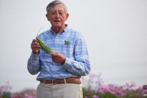 An elderly man in a blue plaid shirt stands outside, holding a bitter melon. Pink flowers and green plants are behind him under clear sky.