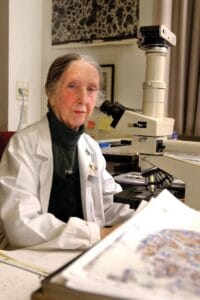 An older woman in a lab coat sits at a desk with a microscope and papers, working in a laboratory or research environment.