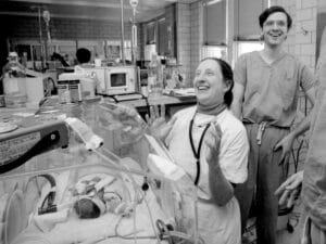 A female doctor smiles and raises her hands in joy near an incubator with a premature baby, as a male doctor in scrubs smiles nearby.