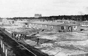 Black and white photo of a World War II prison camp with long, low wooden barracks, groups of people walking on dirt paths, and a forest in the background.