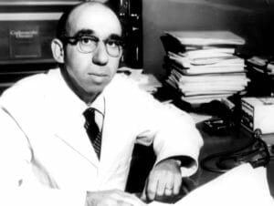 A man in glasses and a white lab coat sits at a paper- and book-covered desk, looking at the camera in a black-and-white photo.