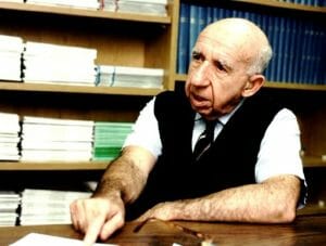 An elderly bald man at a table points at a document. He wears a vest, tie, and short sleeves. Books and papers fill the shelves behind.