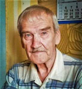 An elderly man with gray hair and a mustache wears a striped shirt and looks at the camera. A wall calendar showing June is visible in the background.