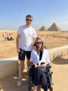 A young man stands beside a young woman in a wheelchair in front of the Pyramids of Giza. The sky is clear and other tourists are visible in the background on the sandy landscape.