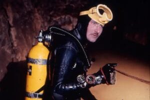 A diver in a black wetsuit and yellow scuba gear, with a yellow tank and goggles, sits in an underwater cave.