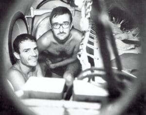 Two shirtless men sit in a cramped, cylindrical space with pipes and machinery. One smiles; the other looks neutral. Black and white photo.
