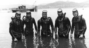 Five divers in vintage scuba gear stand in shallow water, posing with a boat and mountains behind them. The photo is black and white.