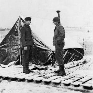 Two soldiers face each other on a snowy path by a snow-covered tent, both in winter uniforms—one American with a helmet, one German with a cap.