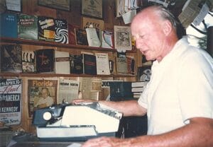 A man in a white shirt uses a typewriter in a room filled with books, magazines, and newspapers covering the walls. He appears focused on his work, surrounded by vintage literature and memorabilia.