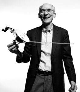 An older man in a suit and glasses smiles, holding a detailed spacecraft model against a plain white background.
