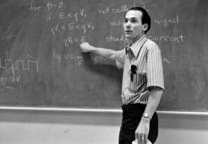 A man in a striped shirt points at math and technical notes on a blackboard.
