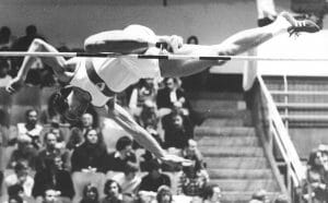 A high jumper soars in mid-air over a horizontal bar at a competition, watched by spectators seated in bleachers behind.