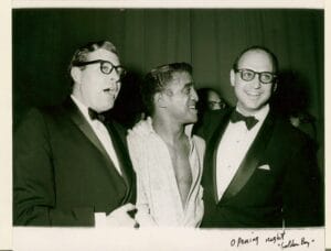 Three smiling men stand together; center in a loosely buttoned shirt, sides in tuxedos and glasses. Dark background. "Opening night.