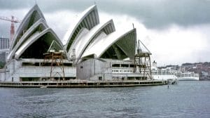 The Sydney Opera House under construction by the waterfront, with cranes and scaffolding visible. Overcast sky and a docked white cruise ship are visible in the background. It is a color photo from the 1960s.
