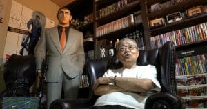 An elderly man with glasses sits in a leather armchair, arms crossed, in a room filled with manga books and a statue behind him.