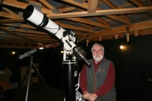 An older man with white hair and a beard stands by a large telescope, under wooden beams. A smaller telescope is in back.
